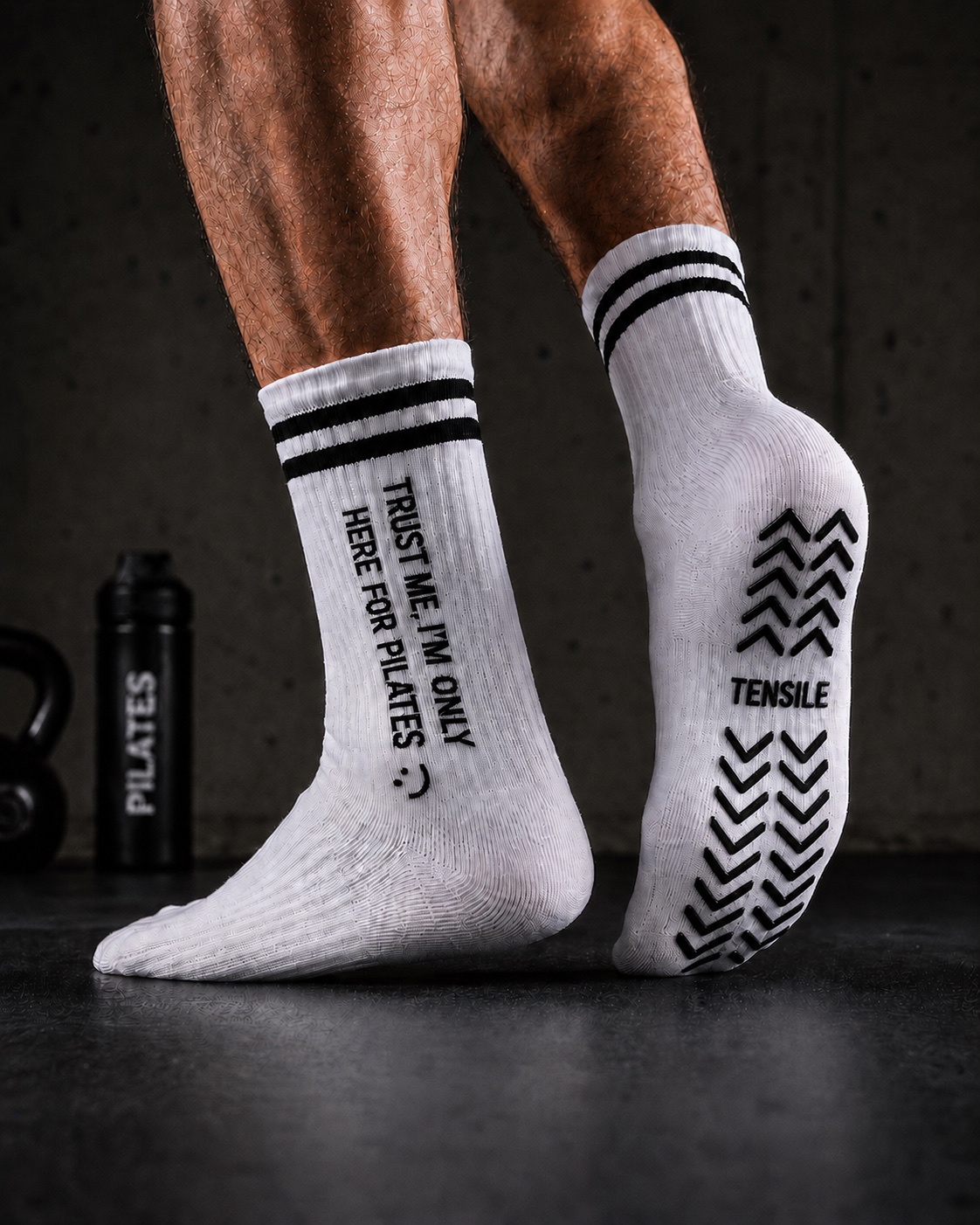 Männerfüße in weißen TENSILE Grip-Socken auf dunklem Gym-Boden, Reformer im Hintergrund